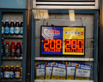 An advertisement display in a store window features lottery jackpot amounts for Mega Millions and Powerball, both showing jackpots of 208 million and 200 million respectively. Below the display, several lottery tickets and promotional posters are visible. To the left of the display, there is a refrigerator stocked with various drinks including different flavors of sports drinks and bottled water.