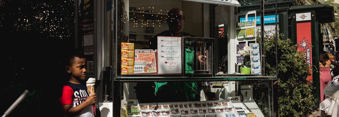 A child holding an ice cream cone stands outside a small kiosk or lottery ticket booth. The booth displays various colorful lottery tickets in the window. There are signs advertising a significant lottery prize amount in Spanish. A person can be seen partially obscured inside the booth, and plants are visible around the area, suggesting an outdoor setting.