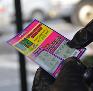 A person wearing black gloves is holding a colorful lottery scratch-off ticket. The ticket includes information about a $1,000,000 guaranteed minimum payout and features multiple games with various prize amounts.